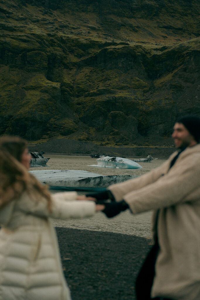 Wide landscape shot of engaged couple standing on vast white glacier in Iceland with mountain peaks in background by Grand Rapids Iceland engagement photographer