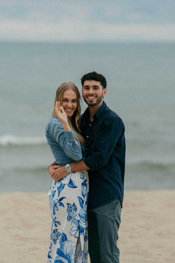 Engaged couple spinning together on Oval Beach shoreline with waves and sunset colors